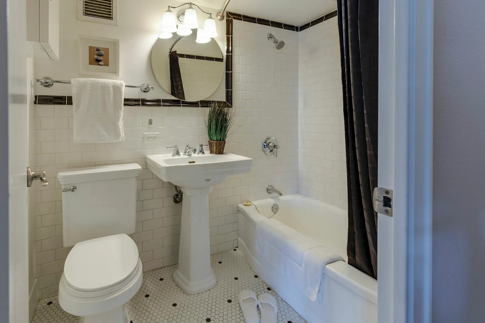 Petite salle de bains en carrelage métro blanc avec frise noire, lavabo sur colonne, miroir rond, baignoire à rideau sombre et sol en tomettes hexagonales