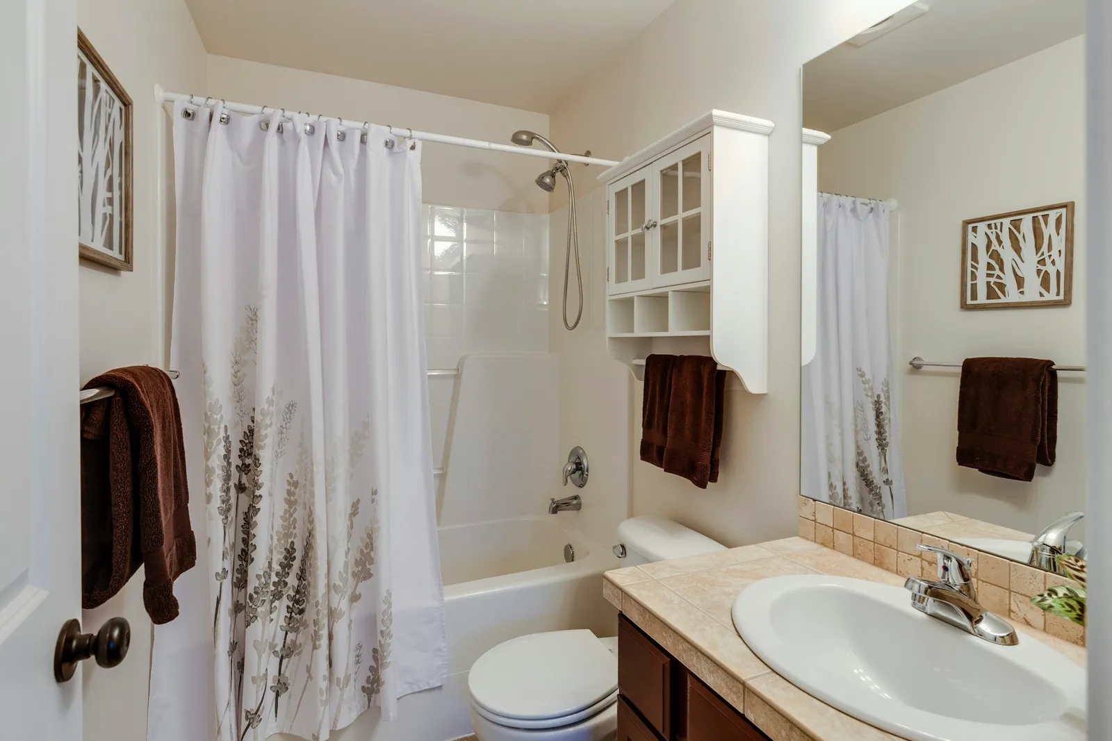 Salle de bains avec plan carrelé beige, vasque ovale, baignoire blanche à rideau fleuri et meuble mural au-dessus du WC