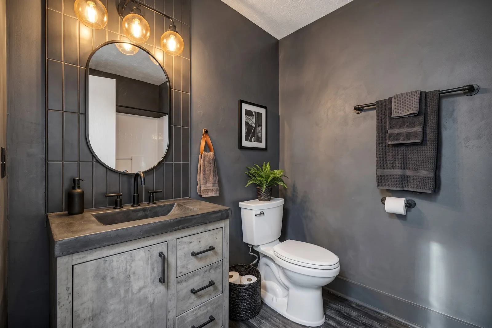 La même pièce en style Loft Industrial avec meuble effet béton, carrelage vertical sombre, miroir ovale noir et ampoules à filament