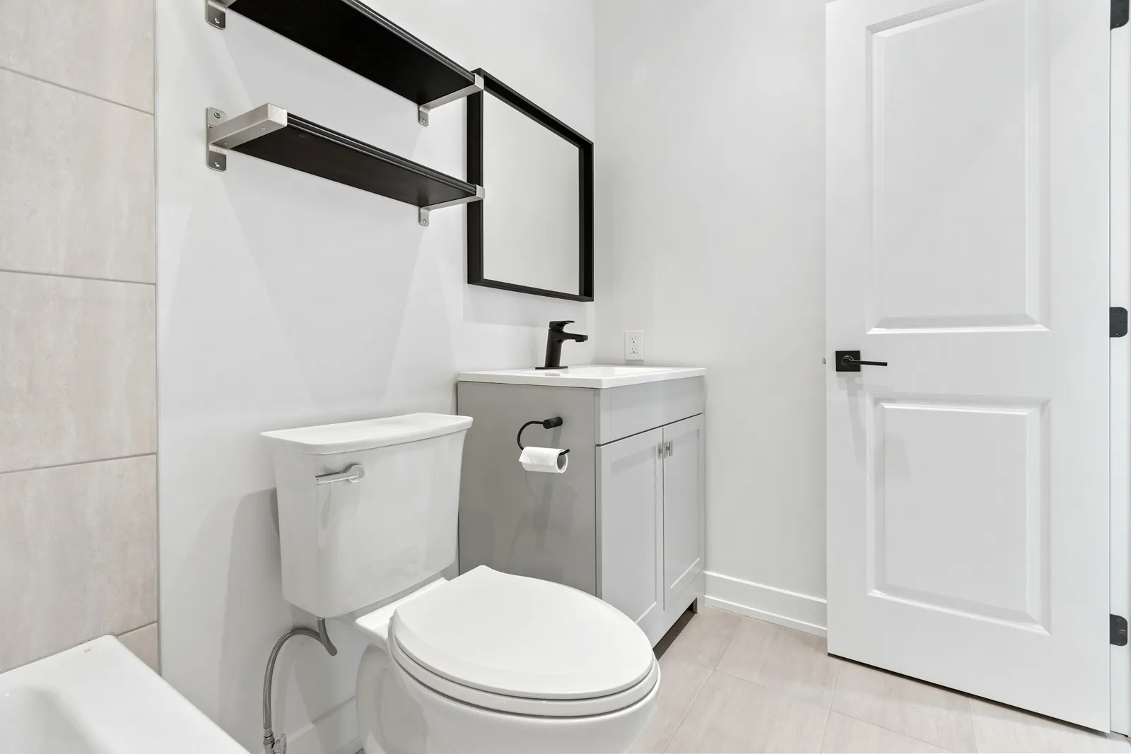 Salle de bains blanche avec meuble vasque gris à cadre, étagères murales noires, miroir à cadre noir, WC et porte moulurée