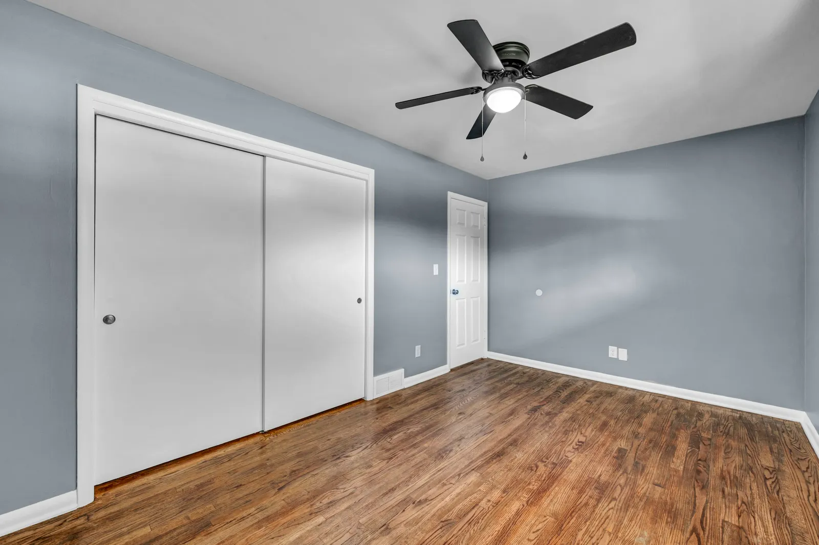 Chambre vide aux murs bleu-gris, placard à portes coulissantes blanches, ventilateur de plafond noir et parquet