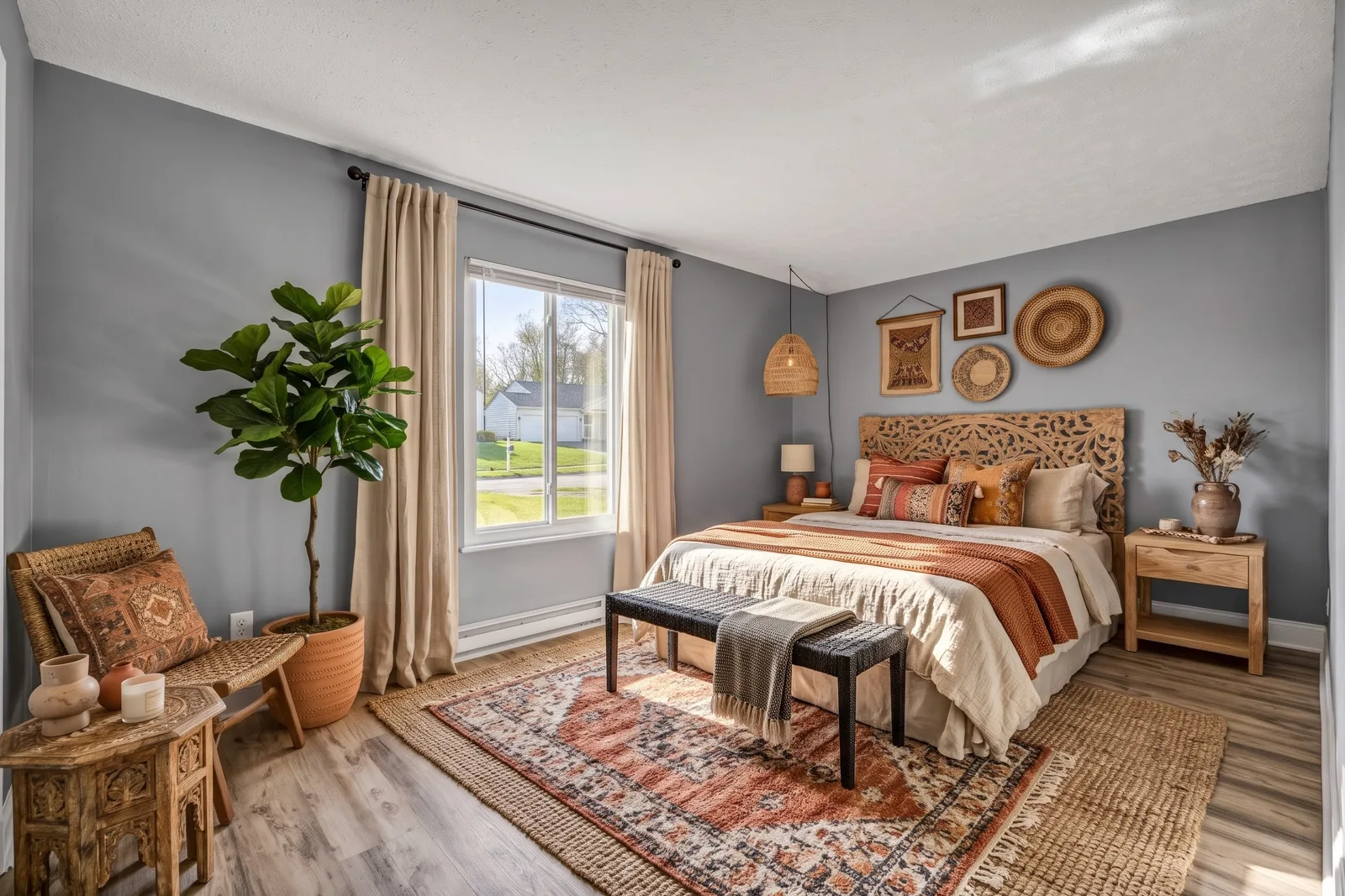 La même chambre en style bohème avec tête de lit sculptée, linge terracotta et crème, tapis à motifs, fauteuil en rotin et ficus lyrata