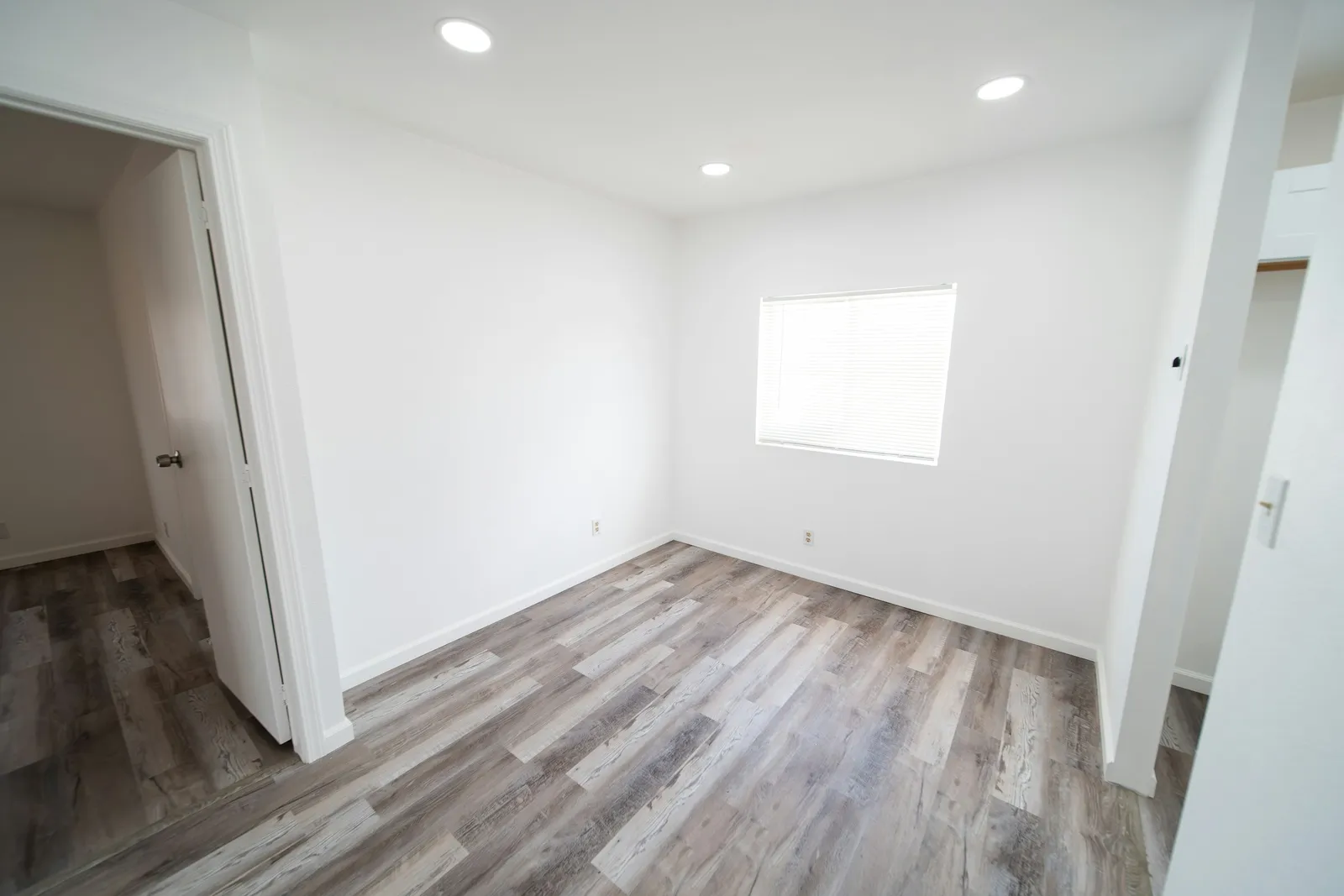 Petite chambre vide vue depuis la porte, murs blancs, fenêtre avec store, spots encastrés et sol vinyle gris