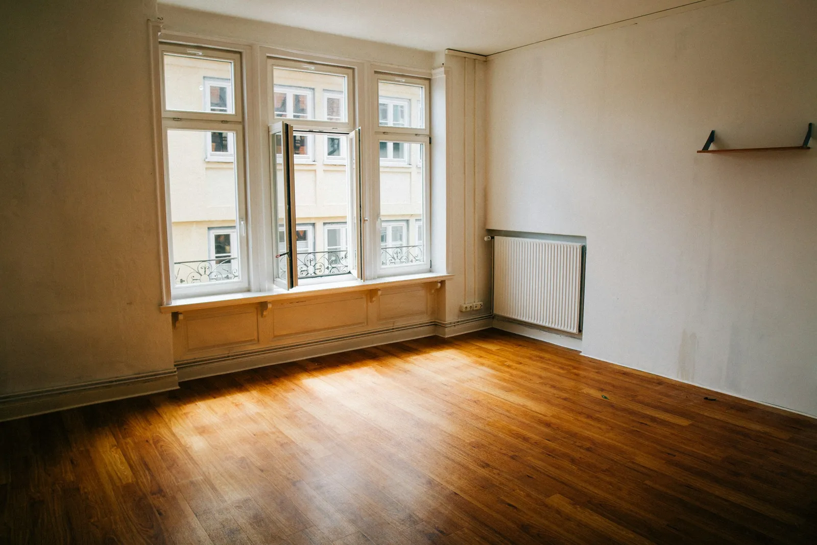 Pièce vide avec hautes fenêtres à petits bois, banquette lambrissée, radiateur, petite étagère murale et parquet