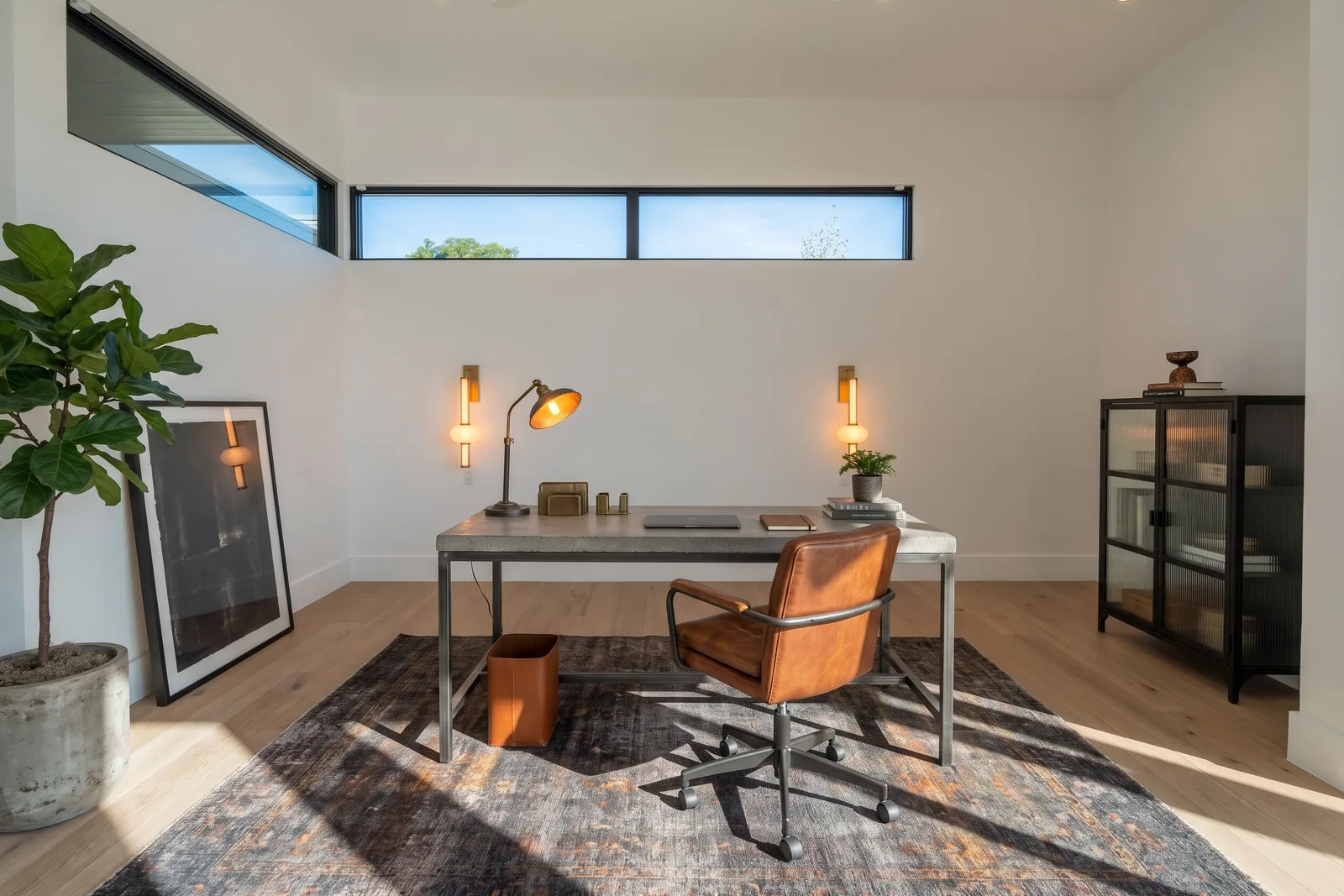 La même pièce en bureau Loft Industrial avec bureau à plateau béton, fauteuil en cuir cognac, vitrine noire, ficus lyrata et tapis sombre