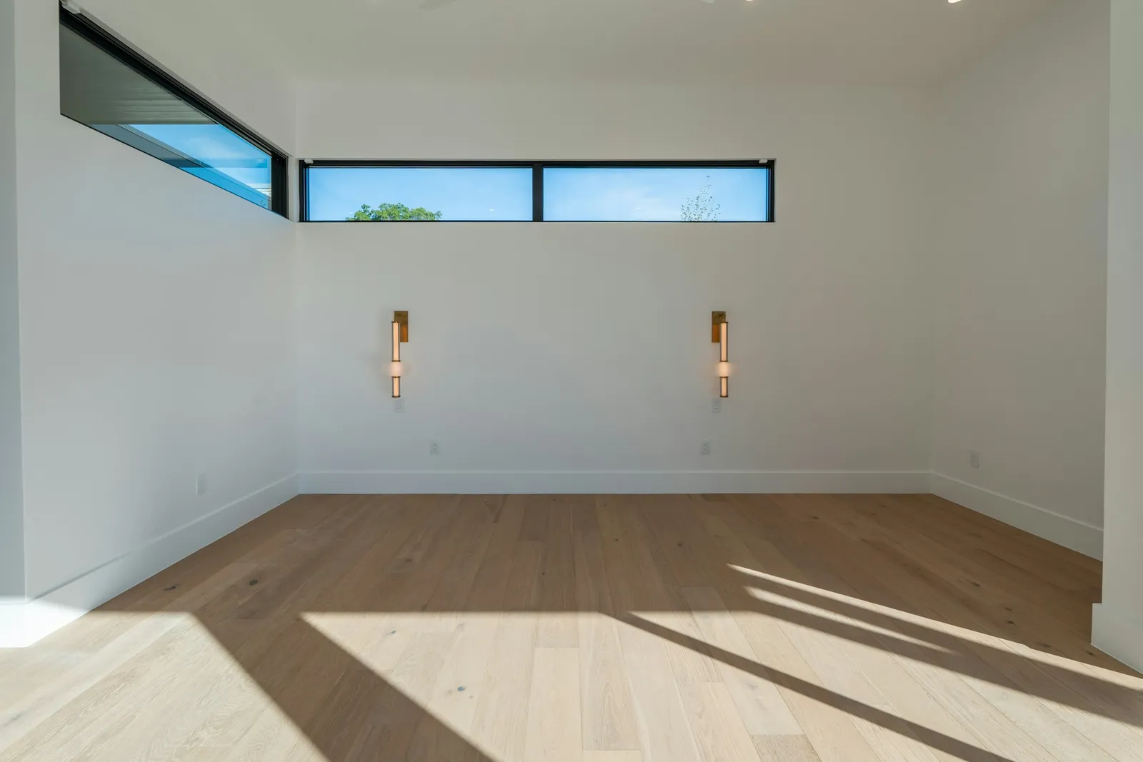 Pièce vide avec fenêtres hautes en bandeau, deux appliques en laiton, murs blancs et parquet en chêne clair