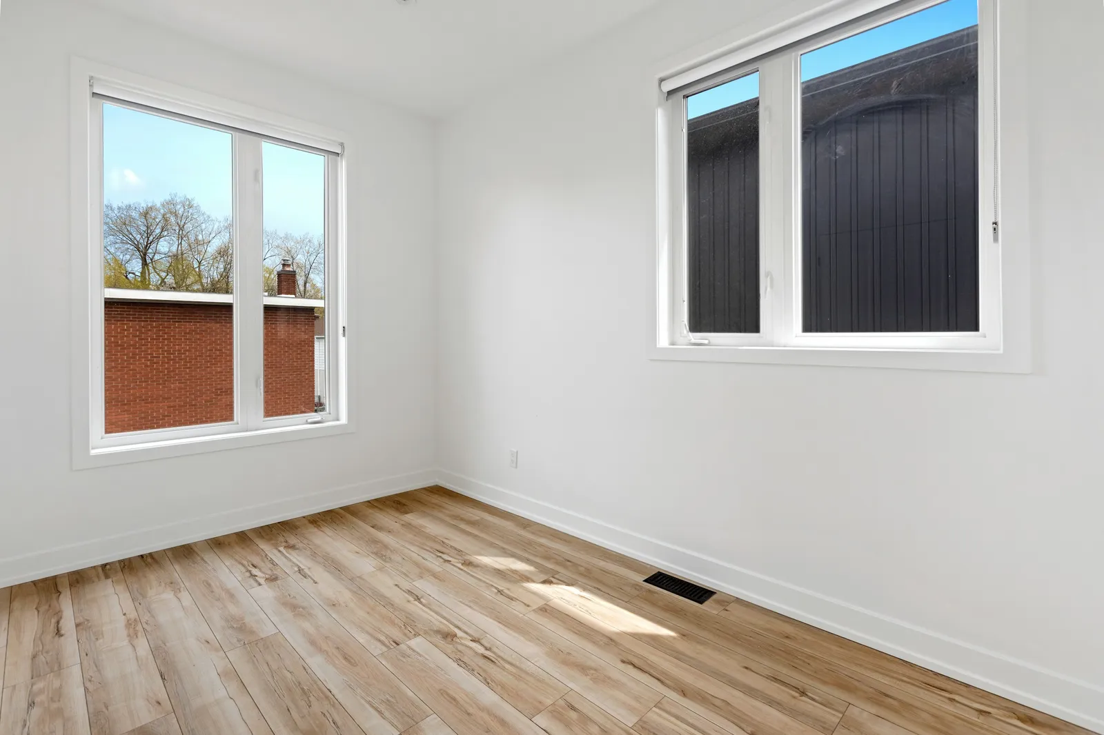 Petite pièce vide avec deux fenêtres à cadre blanc, murs blancs, grille de ventilation au sol et sol imitation bois clair