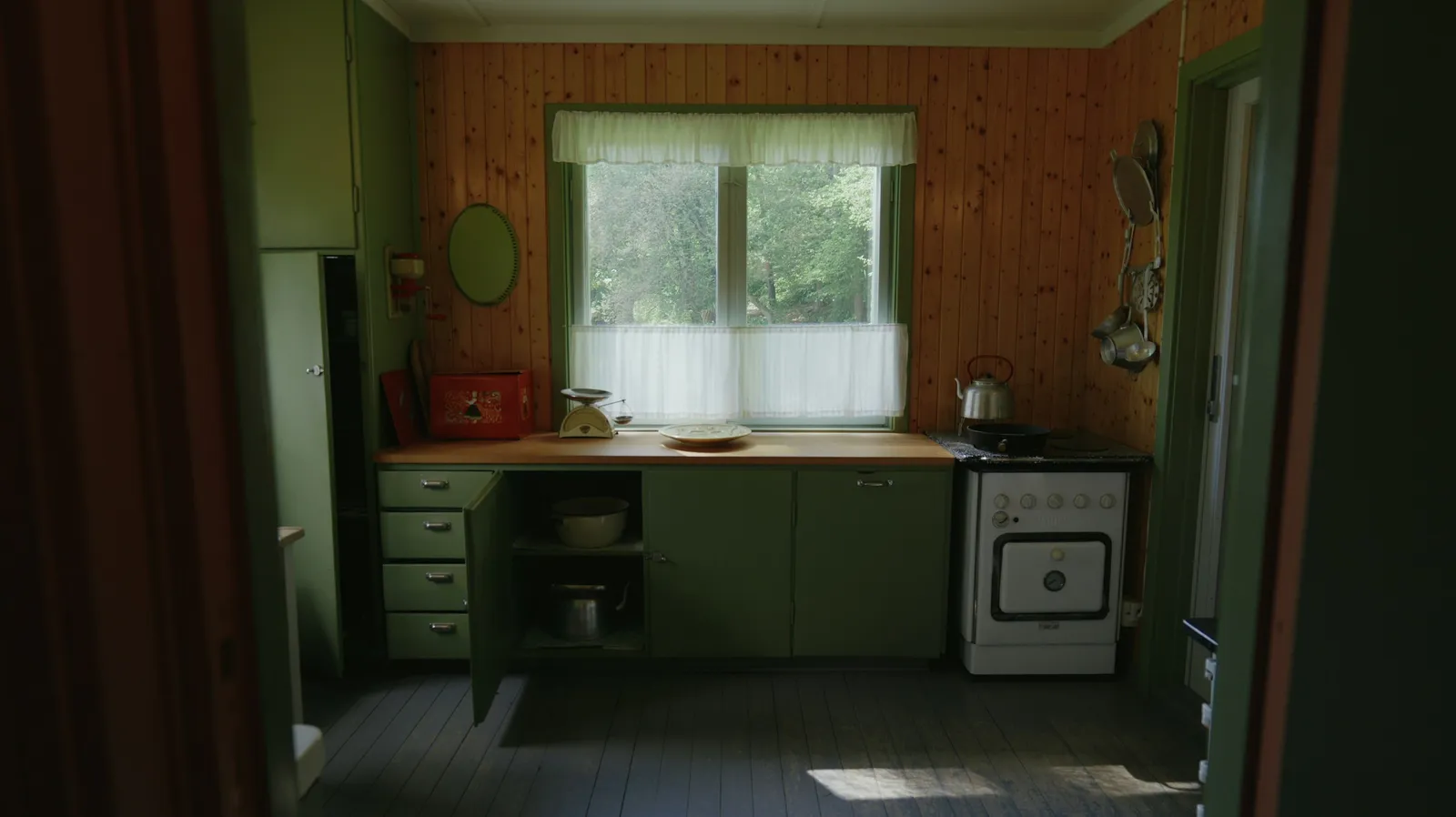 Petite cuisine aux façades vertes, murs lambrissés de pin, cuisinière émaillée blanche et fenêtre à rideaux