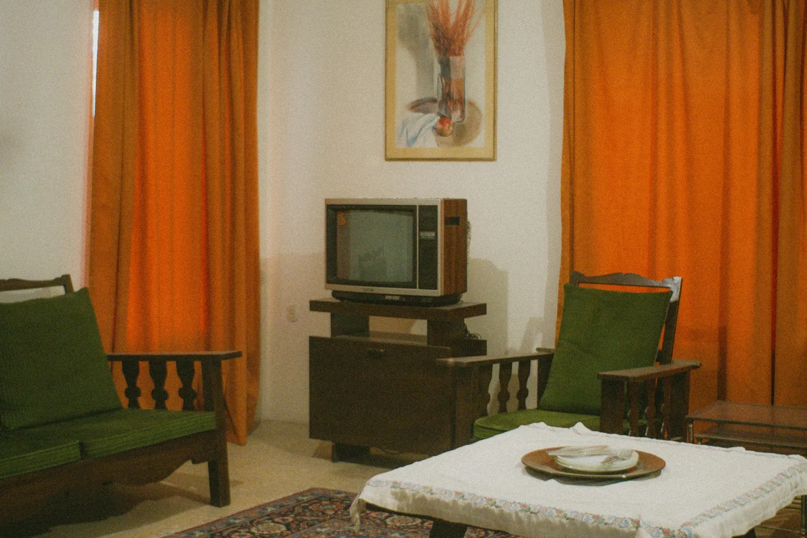 Salon rétro avec rideaux orange, vieux téléviseur sur un meuble en bois, fauteuils verts et table basse couverte d'une nappe