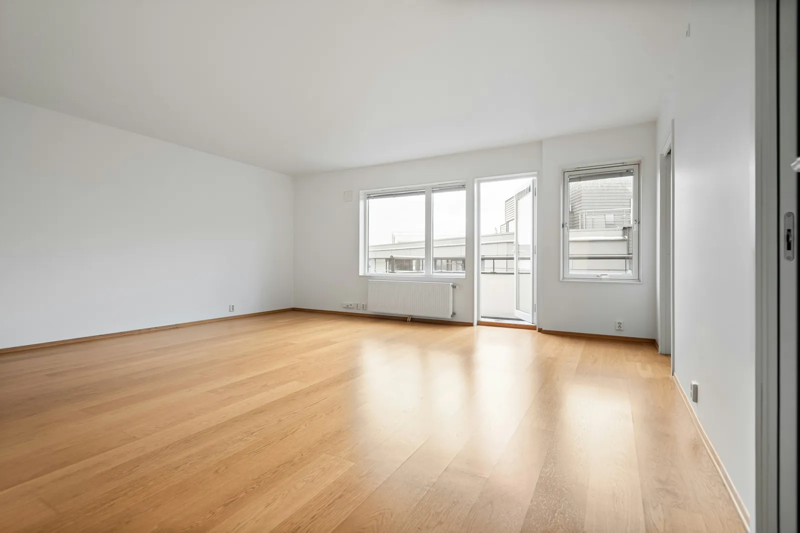 Séjour vide d'appartement avec parquet en chêne, murs blancs, deux fenêtres, une porte de balcon et un radiateur