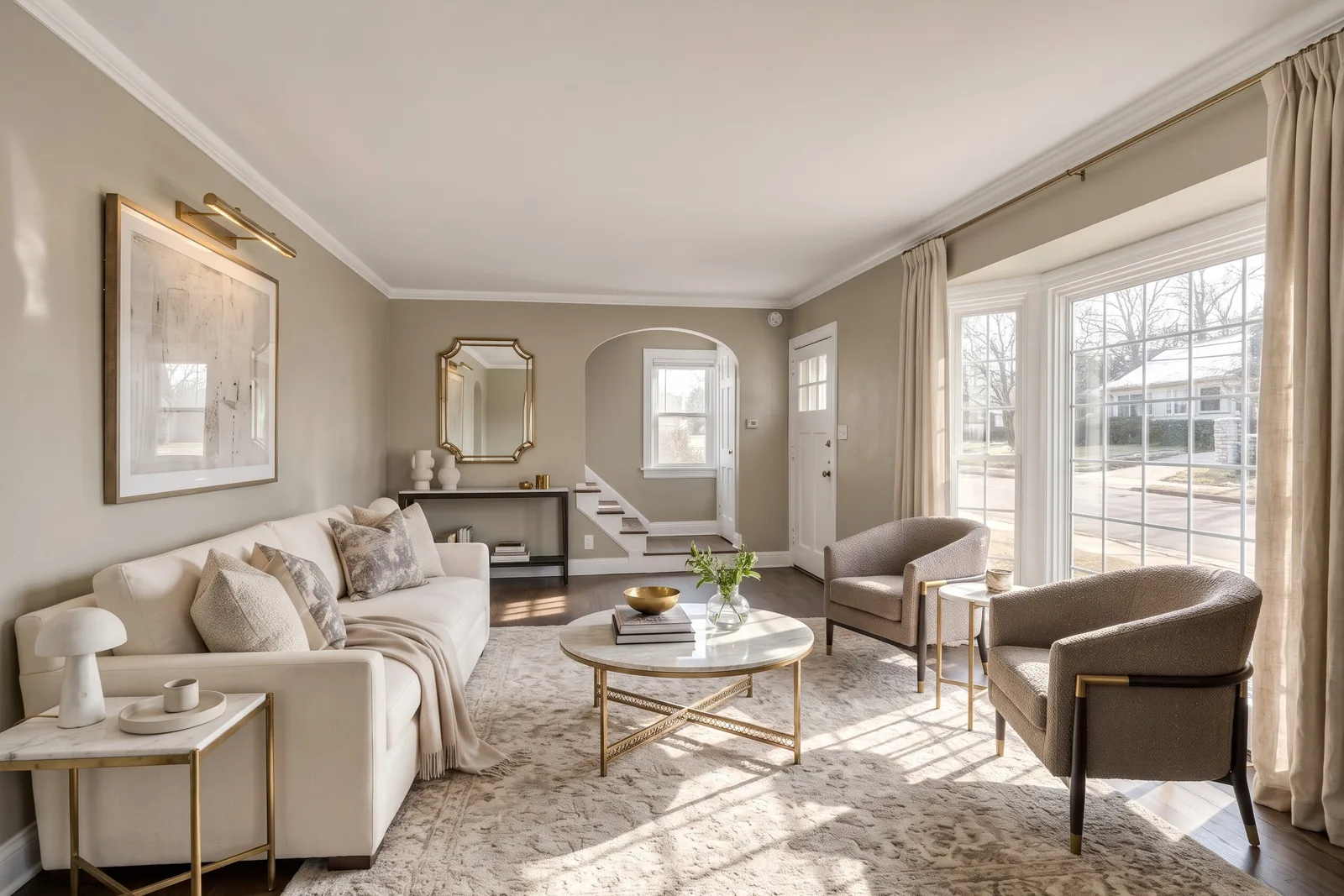 La même pièce meublée en style Transitional Luxe avec canapé crème, deux fauteuils taupe, table basse ronde en marbre et rideaux en lin